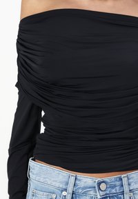 Black off-the-shoulder top with ruched texture, long sleeves, and a fitted design paired with light blue denim jeans.