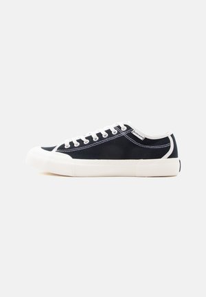 Zwarte lage canvas sneaker met witte rubberen zool, neuskap, veters en stiksels, zijaanzicht tegen een effen achtergrond.