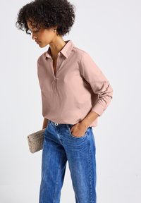 Blusa colletto a piede di pesca con maniche lunghe e scollatura con bottoni, abbinata a jeans in denim blu chiaro e una clutch intrecciata beige.