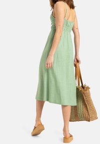 Robe midi en lin vert avec une taille froncée, des bretelles ajustables et une silhouette fluide, associée à un sac fourre-tout tissé marron.