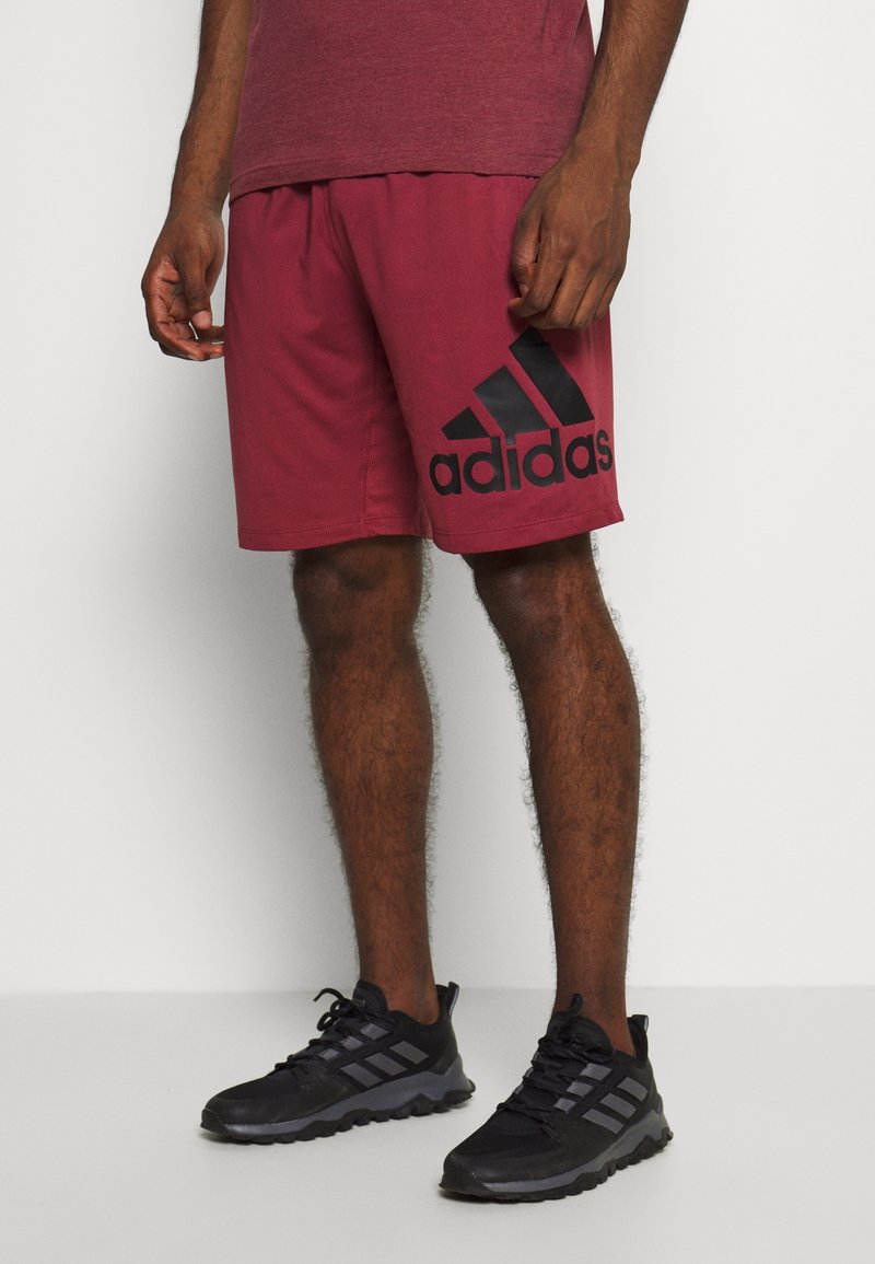 Homme portant un short Adidas bordeaux avec un grand logo et des baskets noires Adidas, debout contre un mur blanc.
