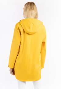 Impermeable amarillo con capucha, fabricado con material resistente al agua. Presenta un corte recto y botones de presión en las mangas. Textura suave y diseño informal.
