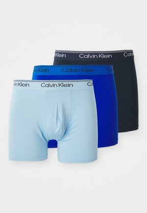 Tres pares de bóxer Calvin Klein en azul claro, azul marino y negro, apilados uno al lado del otro con cinturas de marca.