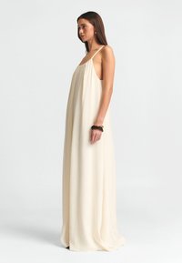 Maxikjole i lys beige stof med tynde stropper, løs pasform og en glat tekstur med en svajende silhuet.