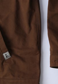 Καφέ υφασμάτινο τζάκετ με τμήμα που περιλαμβάνει μία ορθογώνια ετικέτα με ένα στυλιζαρισμένο γράμμα 'F.' Λείο υλικό, ραμμένες ραφές, χωρίς ορατά σχέδια.