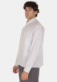 Camicia bianca a maniche lunghe con righe verticali viola e grigie. Caratteristiche distintive includono un colletto con bottoni e maniche con polsini.