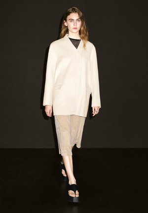 Beige oversize kabát s šálovým límcem, kombinovaný se světlým hnědým vícestupňovým sukní a černými platformovými sandály, na tmavém pozadí.