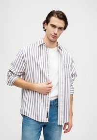 Csíkos ing könnyű anyagból, függőleges lila és kék csíkokkal, gombos elülső része, egyetlen zsebével a mellén és felgöngyölített ujjaival.