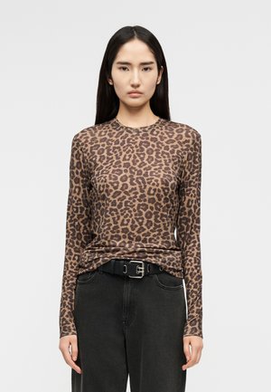 Γυναίκα με ίσια σκούρα μαλλιά φοράει μακρυμάνικο τοπ με καφέ σχέδιο λεοπάρ, το οποίο είναι μπατισμένο μέσα σε μαύρο παντελόνι με μαύρη ζώνη, μπροστά σε απλό φόντο.