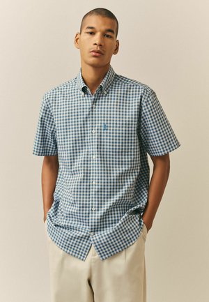 Young man standing with hands in pockets wearing blue and white checkered short-sleeve button-up shirt and light beige pants against plain background.