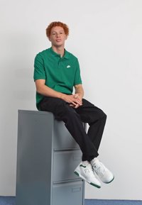 Vihreä Nike-polo paita, mustat housut ja valkoiset New Balance -kengät. Istuu harmaalla arkistokaapilla valkoista taustaa vasten.