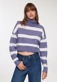 Kobieta w krótkim fioletowo-białym swetrze z golfem w paski i wysokich niebieskich dżinsach, stojąca z jedną ręką w kieszeni.