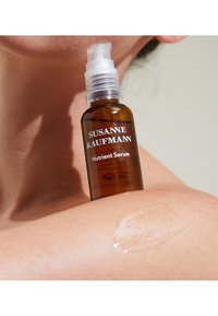 Brązowa szklana butelka z białą pompką, opisana jako „Serum odżywcze Susanne Kaufmann.” Przezroczyste serum widoczne na skórze, matowa powierzchnia.