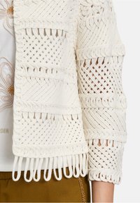 Cardigan tricoté blanc avec un design tissé et des bords à franges. Le tissu a un fini texturé et des manches trois-quarts.
