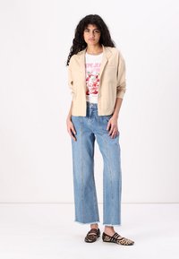 Chaqueta beige de botones sobre una camiseta gráfica blanca, combinada con unos vaqueros de pierna ancha azul claro y unos zapatos planos con estampado negro y beige.