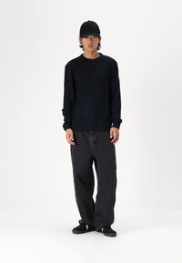 Suéter negro de punto con diseño de cable texturizado, combinado con jeans oscuros sueltos y zapatos negros. El modelo lleva una gorra negra.