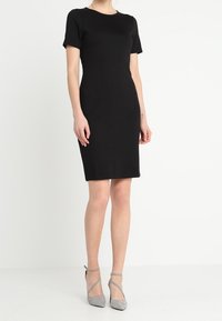 Robe noire ajustée avec des manches courtes, encolure ronde, ourlet à mi-genoux, texture lisse, associée à des chaussures à talons hauts à lanières grises.