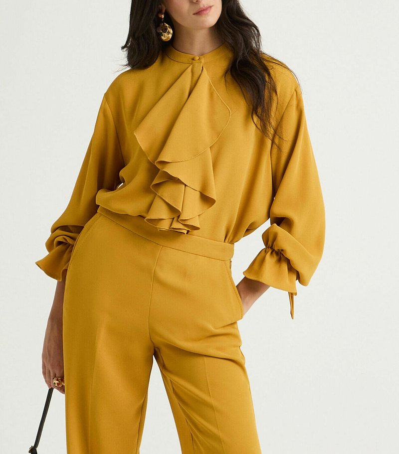 Blusa giallo senape con ampia balza, collo alto e maniche con fiocco. Abbinata a pantaloni sartoriali abbinati.