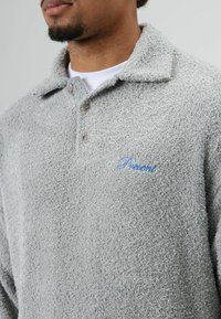 Zachte grijze trui met een kraag, voorzien van bruine knopen en blauwe borduursels met het woord "Present" op de linkerborst. Zachte textuur.