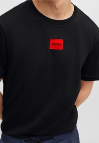 Musta lyhythihainen T-paita pyöreällä pääntiellä, jossa on punainen suorakaiteenmuotoinen kangasmerkki, jossa on mustalla kirjailtu "HUGO", sileä kangaspinta.