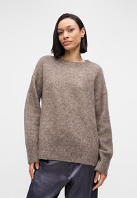 Γυναίκα με φαρδιά, μακριά μανίκια καφέ μάλλινο πουλόβερ και σκούρο σατέν παντελόνι, στέκεται με αυτοπεποίθηση μπροστά σε ένα απλό ανοιχτό γκρι φόντο.