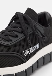 Černé síťové tenisky s kulatou špičkou, plochými šňůrkami a kontrastním bílým logem. Vyznačují se texturovaným svrškem a robustní bílou podrážkou pro pohodlí.