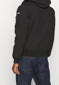 Veste bomber noire avec capuche, poignets élastiques et une poche zippée sur le bras gauche. Fabriquée en matériau lisse et léger.