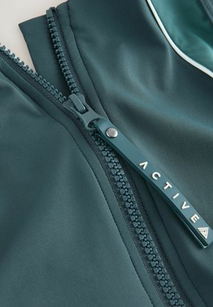 Aktīvās valkāšanas jaka tumši tirkīza krāsā ar gludu tekstūru, kas izceļ kontrastējošu rāvējslēdzēju un logotipa detaļas, uzsverot funkcionālo dizainu.