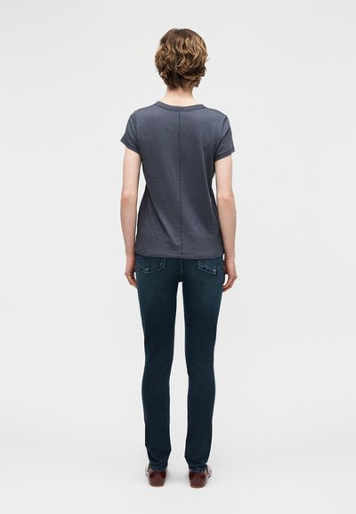 Άτομο με κοντά κυματιστά μαλλιά, που φοράει μια απλή σκούρα γκρι κοντομάνικη μπλούζα, σκούρα μπλε τζιν και καφέ παπούτσια, στέκεται με την πλάτη προς τα εμπρός.