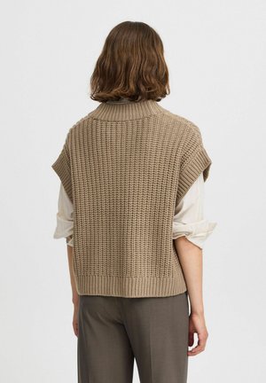 Gilet lavorato a maglia beige con colletto alto e maniche corte, caratterizzato da una trama testurizzata e orlo a costine. Indossato sopra una camicia di colore chiaro.