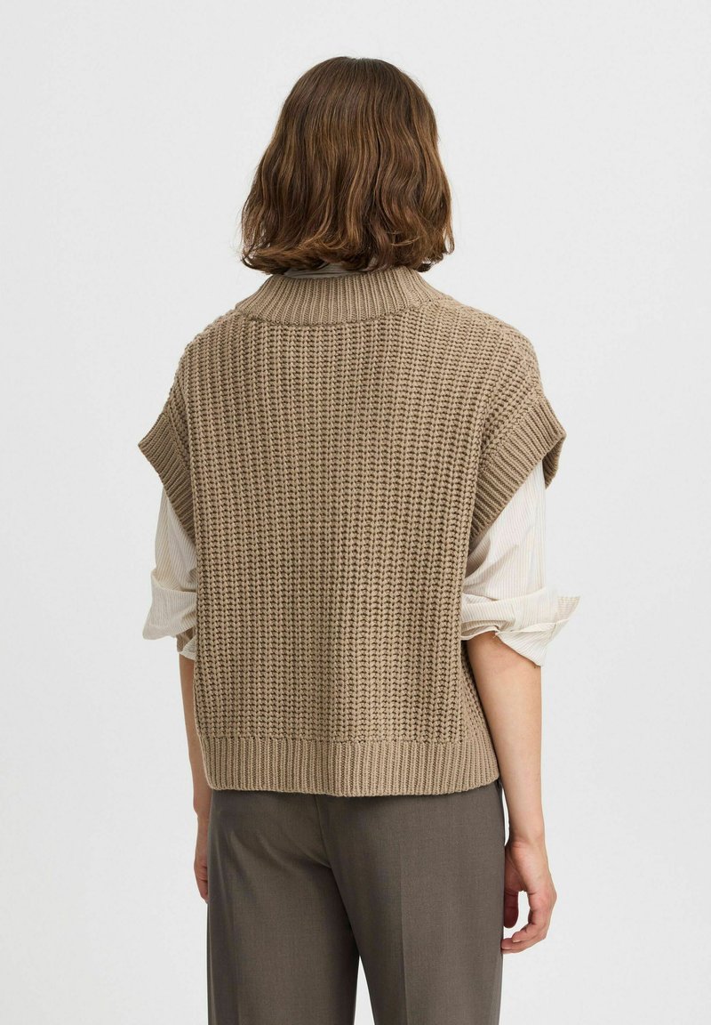 Gilet lavorato a maglia beige con colletto alto e maniche corte, caratterizzato da una trama testurizzata e orlo a costine. Indossato sopra una camicia di colore chiaro.