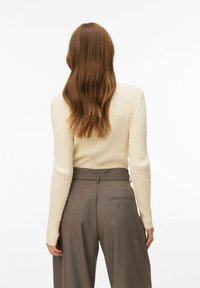 Femme aux cheveux longs ondulés châtain portant un pull côtelé crème et un pantalon large marron taille haute, debout devant un fond blanc.