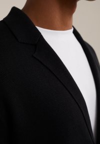 Svart stickad cardigan med slagkrage, figursydd design, som visar en vit t-shirt under. Närbild fångar textur och materialdetaljer.