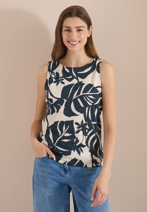 Jeune femme souriante, portant un haut sans manches avec un grand imprimé de feuilles foncées et un jean bleu, debout devant un fond beige uni.