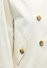 Κοντινό πλάνο μπεζ παλτού με τρία κουμπιά από χελώνα και μια τσέπη με καπάκι στην μπροστινή πλευρά.