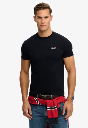 Uomo con T-shirt nera aderente, jeans blu e maglione a righe rosse legato in vita, in piedi con espressione neutra.