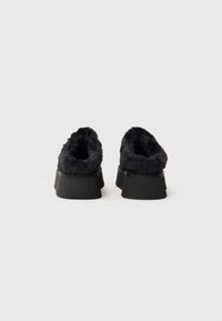 Schwarze plüschige Slipper mit einer flauschigen Textur, abgerundetem Rückenteil und einer dicken schwarzen Gummisohle. Weiche Innenauskleidung um die Öffnung.