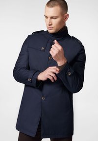 Granatowy płaszcz trench z wysokim kołnierzem, zapinany na guziki, z naszywkami na ramionach oraz wyrazistą fakturą materiału. Posiada skośne kieszenie i guziki na mankietach.