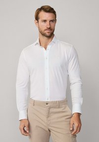 Biała koszula z długim rękawem, zapinana na guziki, o miękkiej teksturze, z pojedynczym logo na klatce piersiowej, w zestawie z jasnobeżowymi spodniami.