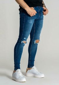 Úzké modré džínové kalhoty s roztrhanými dírami na kolenou, s přiléhavým střihem a klasickým stylem s pěti kapsami, doplněné bílými teniskami.