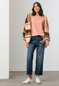 Rosa Pullover mit besticktem Logo, gestreifte oversized Strickjacke in mehreren Farben, blaue gerade Jeans und weiße Sneakers.