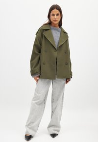 Olive zaļas pārāk liela izmēra divrindu mētelis ar iedziļinātu apkakli, lieliem pogām un sānu kabatām, apvienots ar pelēku džemperi un platu kāju džinsiem.