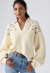 Pull en maille couleur crème avec un col en V, des détails en dentelle aux épaules et une texture côtelée. Associé à un jean bleu et une ceinture marron.