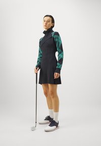 Golf obleka v mornarsko modri barvi z visokim ovratnikom in vzorčenimi teal rokavi; drži golf palico, nosi bele nogavice in dvobarvne golf čevlje.