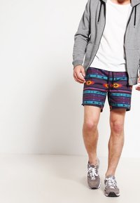 Sudadera gris con cremallera, camiseta blanca y pantalones cortos con estampado en púrpura, azul y naranja con diseños geométricos, combinados con zapatillas deportivas grises.