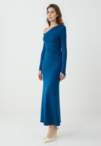 Robe de soirée asymétrique à une épaule, à manches longues, en teal avec des détails froncés et une coupe longue jusqu'au sol ; elle est fabriquée dans un tissu lisse et extensible.