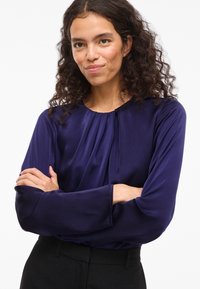 Kvinna med lockigt mörkt hår, iklädd en marinblå blus och svarta byxor, står med armarna i kors mot en vit bakgrund.