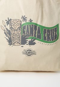 Beige vászontáska, amelyen egy tiki arc grafika található zöld és fekete "SANTA CRUZ" felirattal, alatta pedig a "Roskopp's Voodooroom" logó.
