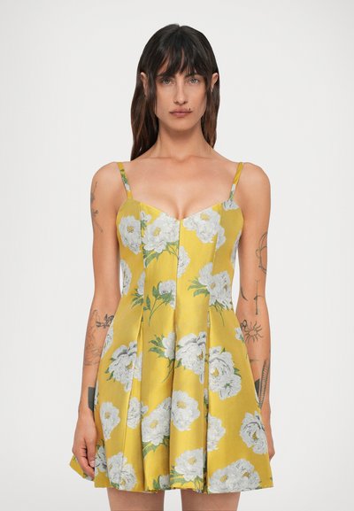 Femme aux longs cheveux noirs portant une robe sans manches jaune à fleurs avec des fleurs blanches, se tenant devant un fond clair uni.