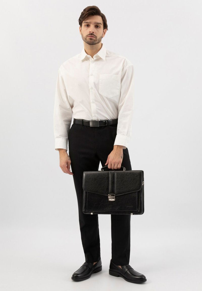 Homme en chemise blanche et pantalon noir tenant une mallette noire, debout devant un fond clair uni.